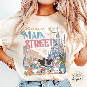 May include: Cream-colored sweatshirt featuring the text "Meet me on Main Street" above a graphic of Disney characters and Cinderella's castle. The design includes Mickey and Minnie Mouse, Donald and Daisy Duck, and Goofy.