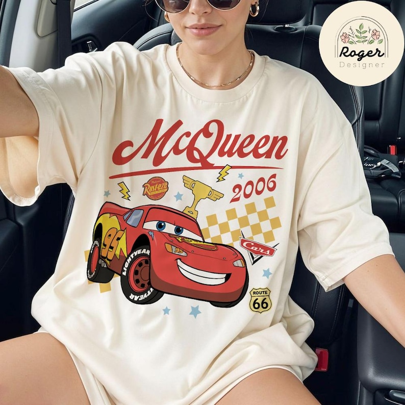 May include: Cream-colored t-shirt featuring a graphic of Lightning McQueen from the movie Cars. The design includes the text "McQueen 2006" in red, a checkered flag, and Route 66 signage. The shirt has a relaxed fit.