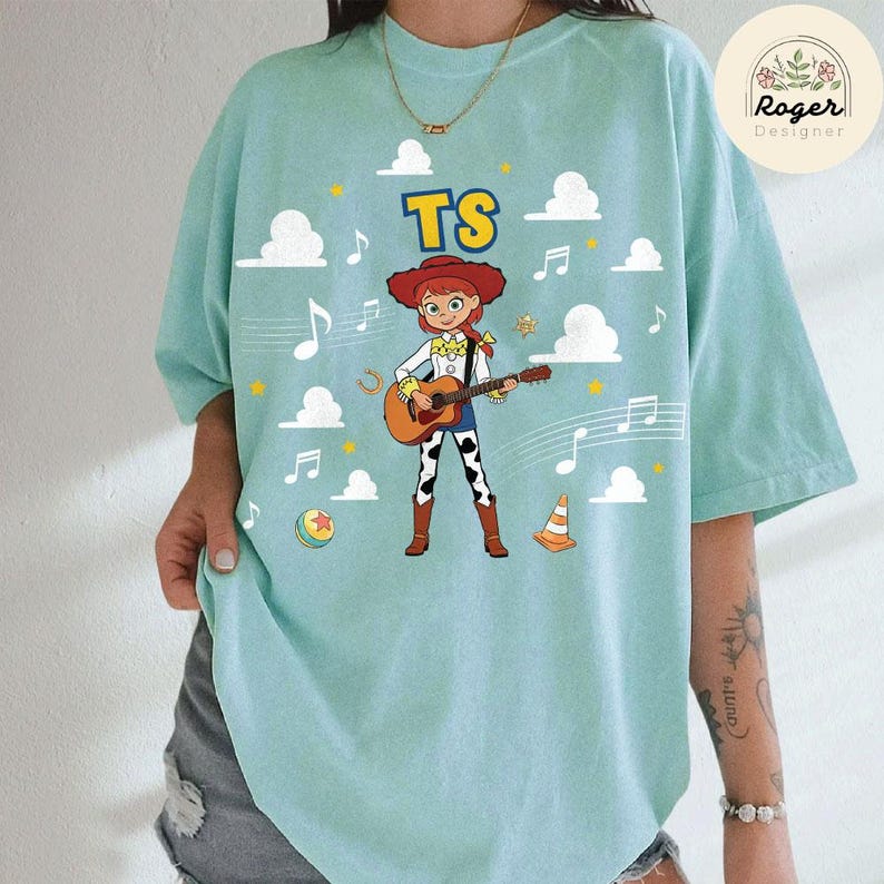 K&ouml;nnte beinhalten: Ein helles mintgr&uuml;nes T-Shirt mit einer Grafik einer Comic-Cowgirl-Figur, die eine akustische Gitarre spielt. Das Design enth&auml;lt den Text TS, wei&szlig;e Wolken, Musiknoten, einen bunten Ball und einen orangefarbenen Verkehrskegel im Hintergrund.
