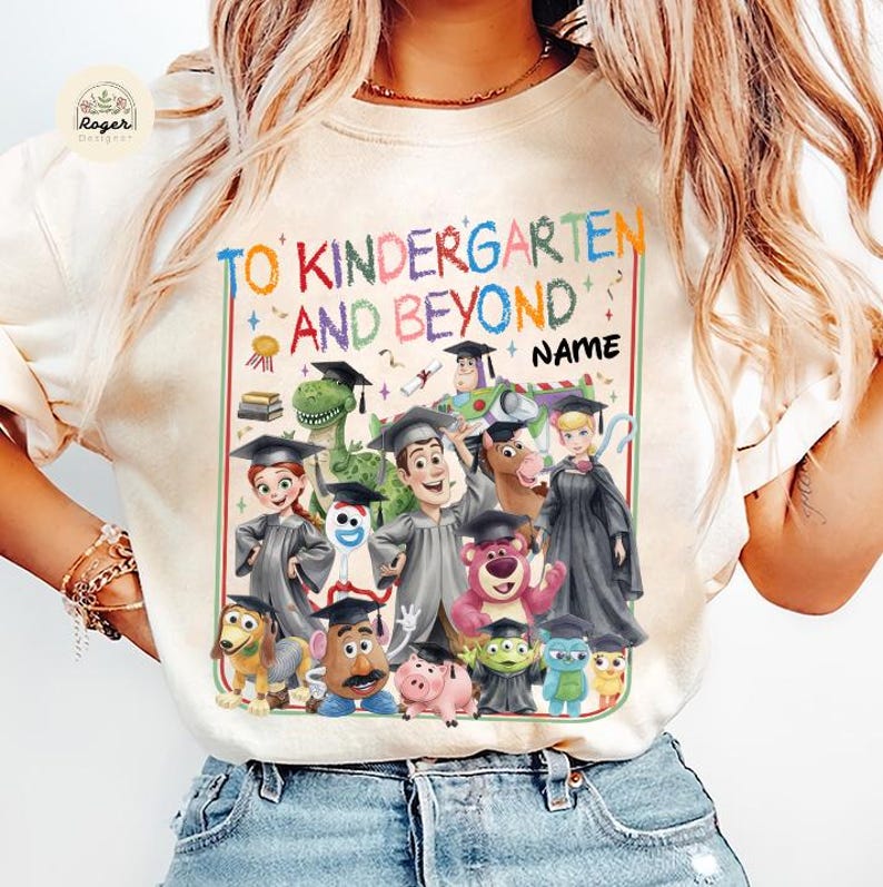 Peut inclure: T-shirt cr&egrave;me avec l'inscription "TO KINDERGARTEN AND BEYOND NAME" en lettres color&eacute;es. Le t-shirt pr&eacute;sente un graphisme de personnages de dessins anim&eacute;s portant des chapeaux et des robes de fin d'&eacute;tudes, parfait pour une remise de dipl&ocirc;me de maternelle.