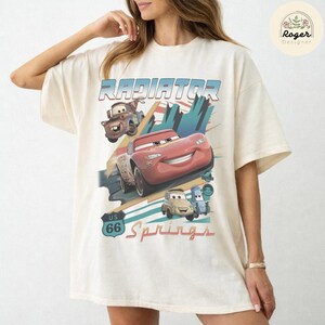 May include: An off-white short-sleeve t-shirt featuring a graphic print of characters from the movie Cars, including Lightning McQueen and Mater. The design includes the text Radiator Springs and a US 66 road sign graphic in blue and red.