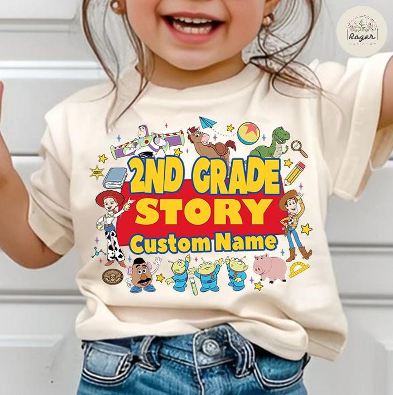 Pu&ograve; includere: Una maglietta color crema con un design grafico colorato. Il design include il testo "2ND GRADE STORY" in giallo e rosso, circondato da personaggi dei cartoni animati. Sono visibili anche le parole "Custom Name".