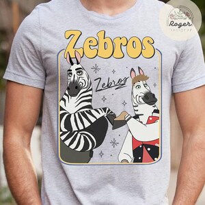 Pu&ograve; includere: T-shirt grigia con una grafica a cartoni animati di due zebre. La parola "Zebros" &egrave; in lettere gialle a bolle sopra le zebre. Le zebre sono bianche e nere con caratteristiche da cartone animato. La maglietta ha uno scollo tondo.