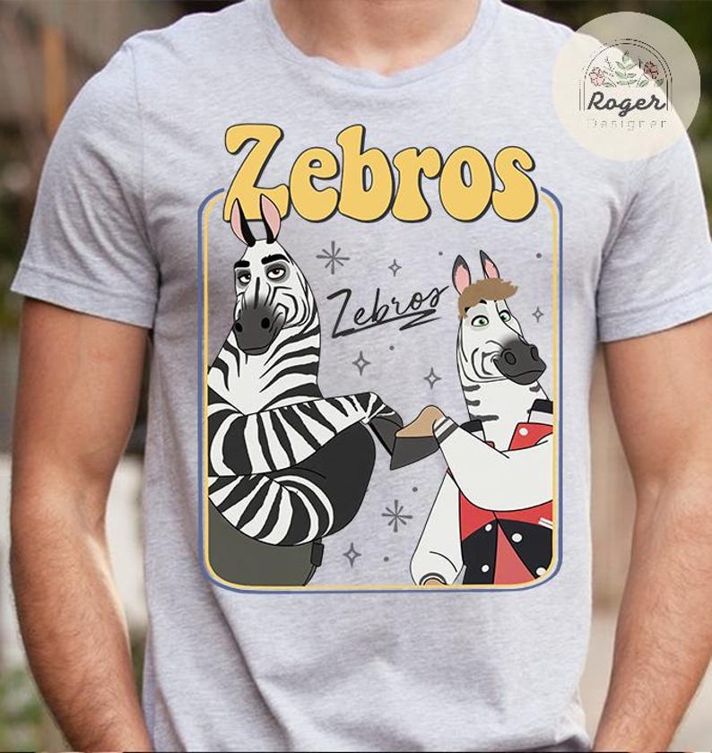 Pu&ograve; includere: T-shirt grigia con una grafica a cartoni animati di due zebre. La parola "Zebros" &egrave; in lettere gialle a bolle sopra le zebre. Le zebre sono bianche e nere con caratteristiche da cartone animato. La maglietta ha uno scollo tondo.