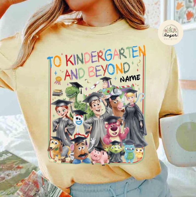 Peut inclure: T-shirt jaune clair avec un motif color&eacute;. Le motif comprend le texte "TO KINDERGARTEN AND BEYOND" et des personnages de dessins anim&eacute;s. Il y a un espace pour un nom. Le t-shirt a un col rond.