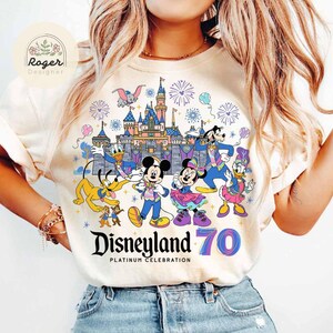 K&ouml;nnte beinhalten: Ein wei&szlig;es T-Shirt mit einer bunten Grafik von Mickey Mouse, Minnie Mouse, Donald Duck, Goofy und anderen Disney-Charakteren, die das 70-j&auml;hrige Jubil&auml;um von Disneyland feiern. Die Grafik enth&auml;lt den Text "Disneyland 70 Platinum Celebration".