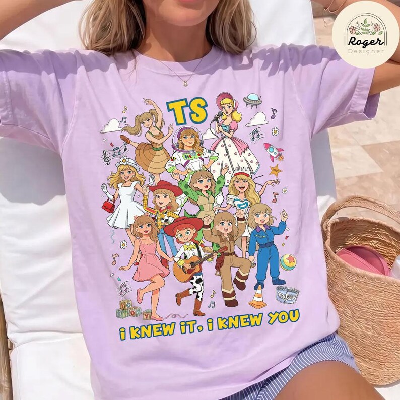 Op de afbeelding: Een lichtpaars t-shirt met korte mouwen en een kleurrijke grafische print van meerdere stripfiguren in verschillende outfits. De tekst TS staat bovenaan en de tekst I KNEW IT, I KNEW YOU staat onderaan de afbeelding gedrukt.