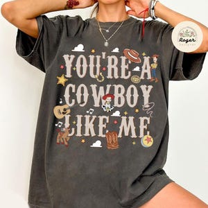 Może przedstawiać: Ciemnoszary t-shirt z kr&oacute;tkim rękawem z napisem "YOU'RE A COWBOY LIKE ME" czcionką w stylu western. Grafika zawiera ilustracje kowbojskiego kapelusza, gitary, lassa, kowbojskich but&oacute;w, piłki oraz postaci z kresk&oacute;wek.