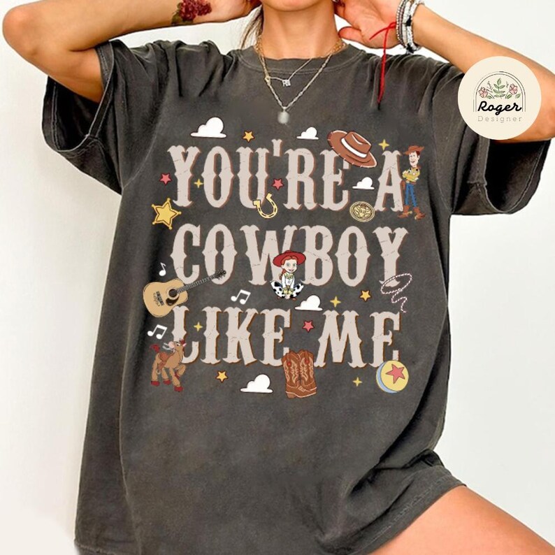 Może przedstawiać: Ciemnoszary t-shirt z kr&oacute;tkim rękawem z napisem "YOU'RE A COWBOY LIKE ME" czcionką w stylu western. Grafika zawiera ilustracje kowbojskiego kapelusza, gitary, lassa, kowbojskich but&oacute;w, piłki oraz postaci z kresk&oacute;wek.