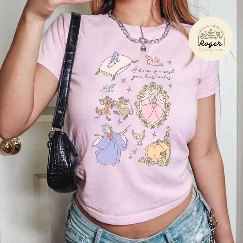 Może przedstawiać: Jasnor&oacute;żowa koszulka z grafiką w stylu Kopciuszka. Projekt zawiera ilustracje Kopciuszka, myszy, dyniowej karety oraz napis "A dream is a wish your heart makes."