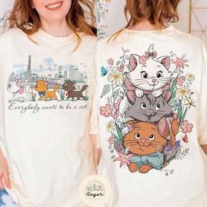 May include: A white t-shirt with a graphic print of three cartoon cats in front of a cityscape. The text "Everybody wants to be a cat" is printed below the cats. The cats are stacked on top of each other, with the top cat being white, the middle cat being gray, and the bottom cat being orange. The cats are surrounded by flowers and butterflies.