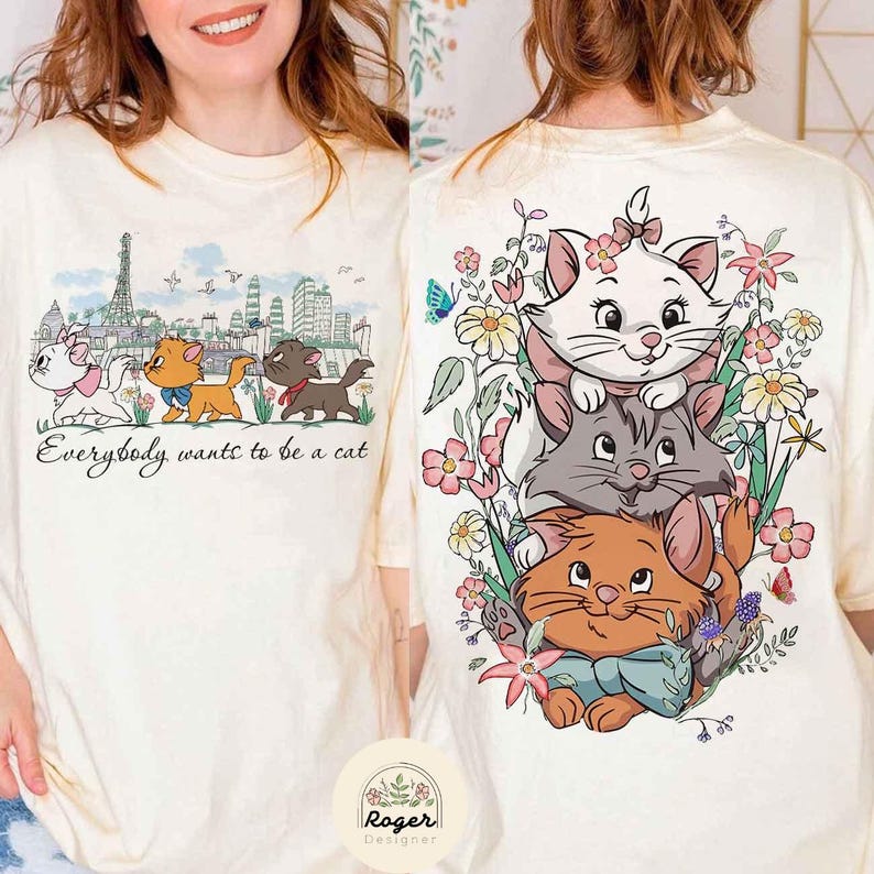 May include: A white t-shirt with a graphic print of three cartoon cats in front of a cityscape. The text "Everybody wants to be a cat" is printed below the cats. The cats are stacked on top of each other, with the top cat being white, the middle cat being gray, and the bottom cat being orange. The cats are surrounded by flowers and butterflies.