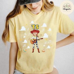 K&ouml;nnte beinhalten: Ein blassgelbes T-Shirt mit einer Grafik einer Comic-Cowgirl-Figur mit roten Haaren, die eine akustische Gitarre spielt. Das Design enth&auml;lt den Text TS, wei&szlig;e Wolken, Noten, einen kleinen orangefarbenen Verkehrskegel und einen bunten Ball.