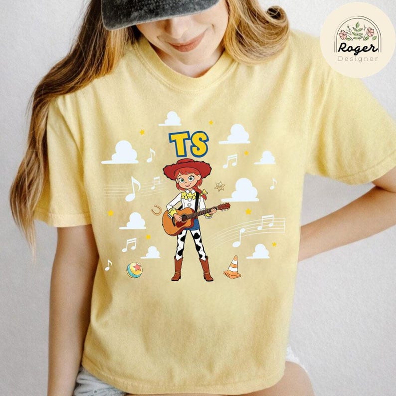 K&ouml;nnte beinhalten: Ein blassgelbes T-Shirt mit einer Grafik einer Comic-Cowgirl-Figur mit roten Haaren, die eine akustische Gitarre spielt. Das Design enth&auml;lt den Text TS, wei&szlig;e Wolken, Noten, einen kleinen orangefarbenen Verkehrskegel und einen bunten Ball.