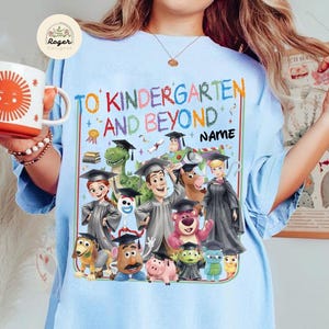 Peut inclure: T-shirt bleu clair avec un motif color&eacute; affichant le texte "TO KINDERGARTEN AND BEYOND NAME". Le motif comprend des personnages de dessins anim&eacute;s en tenue de remise de dipl&ocirc;mes. Une tasse blanche avec un soleil orange est tenue dans l'image.