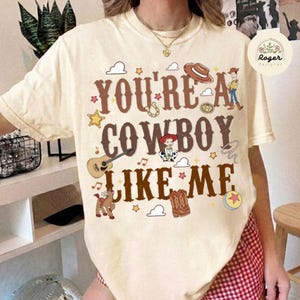 Może przedstawiać: Kremowy t-shirt z kr&oacute;tkim rękawem z brązowym napisem "YOU'RE A COWBOY LIKE ME", otoczony ilustracjami kapelusza kowbojskiego, gitary, lassa, konia, kowbojek, gwiazd, chmur oraz postaciami z serii film&oacute;w Toy Story.