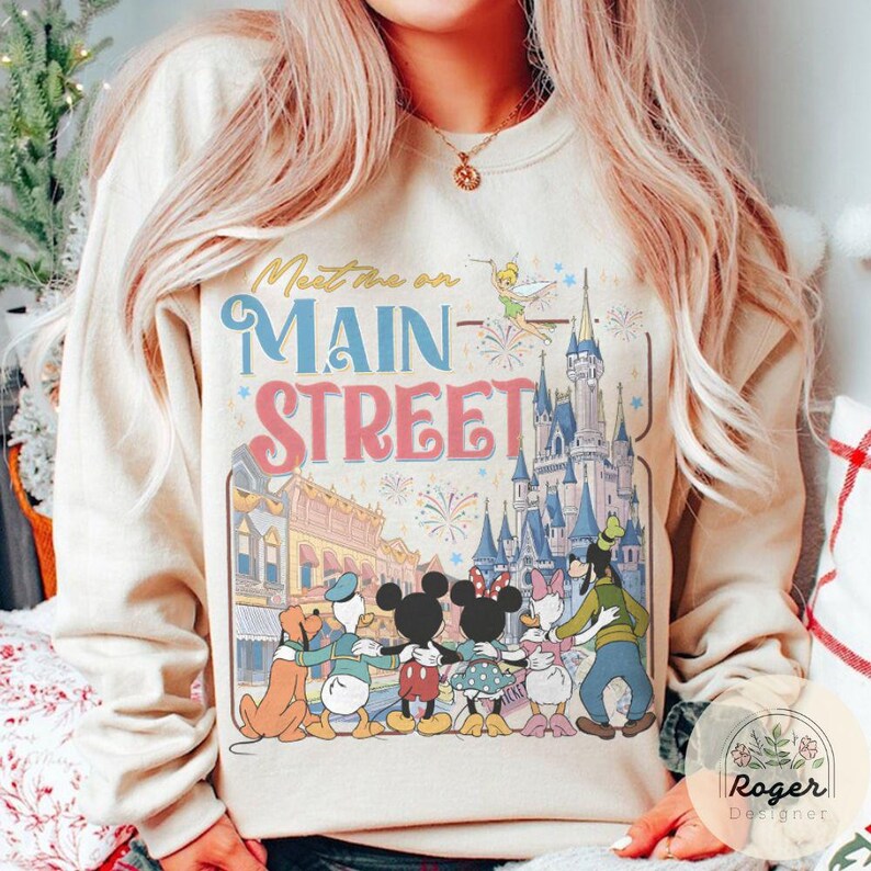 May include: Beige sweatshirt with the text "Meet Me on Main Street" in blue and pink. The design features Disney characters like Mickey and Minnie Mouse, and a castle. The sweatshirt has a crew neck and long sleeves.