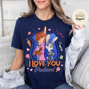 Pu&ograve; includere: T-shirt blu navy con un'illustrazione a cartoni animati di Nick Wilde e Judy Hopps di Zootropolis, con la scritta "I Love You Partner!". Il design include cuori, impronte di zampe e una carota. La maglietta &egrave; girocollo.