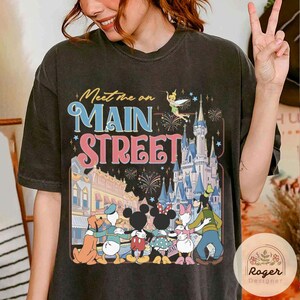 May include: Dark gray t-shirt featuring the text "Meet Me on Main Street" with a colorful graphic of Disney characters, a castle, and fireworks. The design includes Mickey and Minnie Mouse, Donald Duck, Daisy Duck, and Goofy.