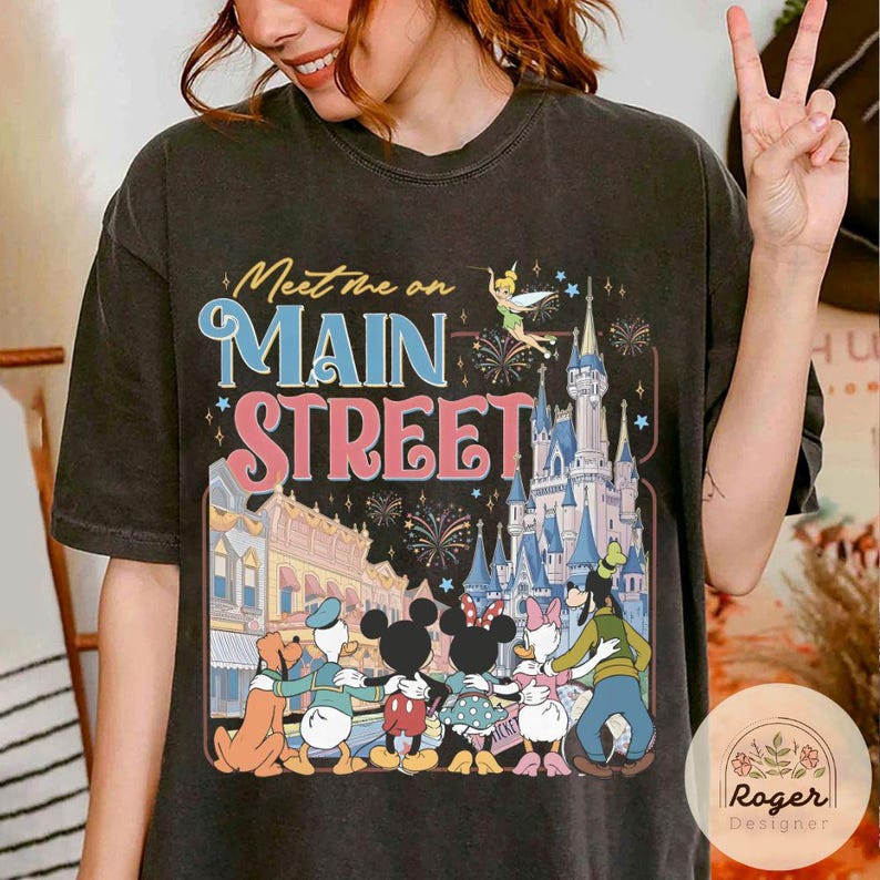 May include: Dark gray t-shirt featuring the text "Meet Me on Main Street" with a colorful graphic of Disney characters, a castle, and fireworks. The design includes Mickey and Minnie Mouse, Donald Duck, Daisy Duck, and Goofy.