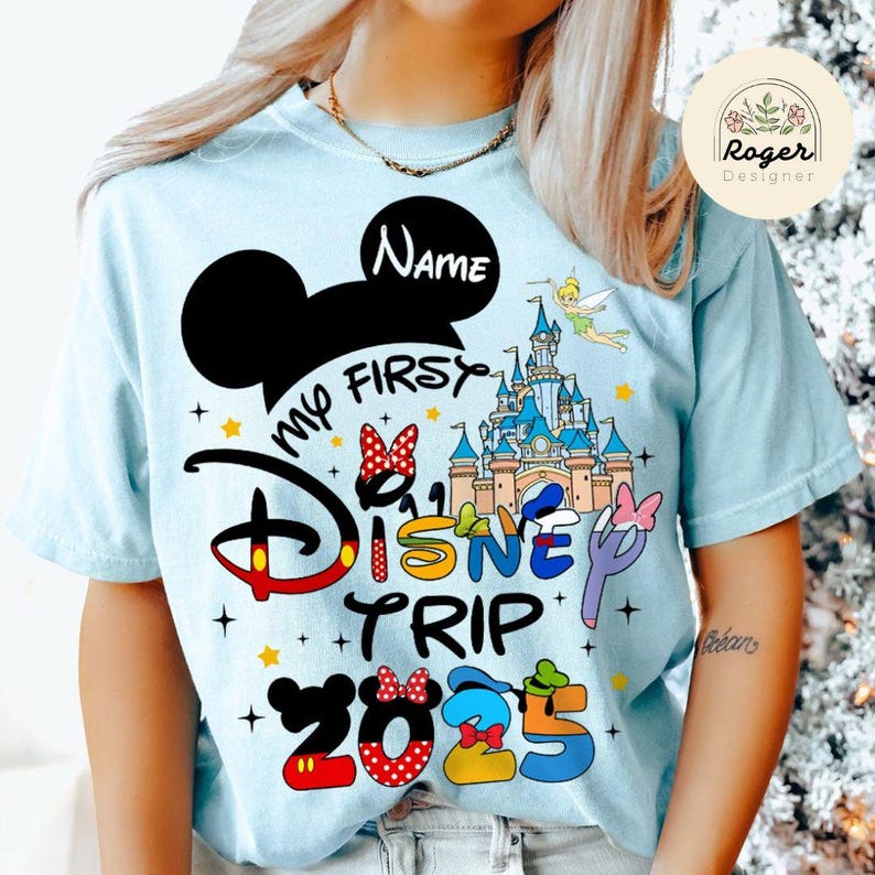 May include: Light blue t-shirt featuring a Mickey Mouse ear design and the text "My First Disney Trip 2025". The shirt also includes a graphic of Cinderella's castle and other Disney characters.