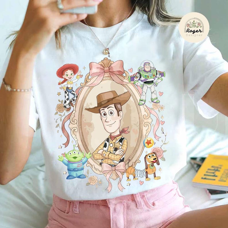 May include: White t-shirt with a cartoon illustration of Woody from Toy Story in a decorative frame, surrounded by characters like Buzz Lightyear and Jessie. The design includes pink bows and floral accents, creating a playful and whimsical aesthetic.