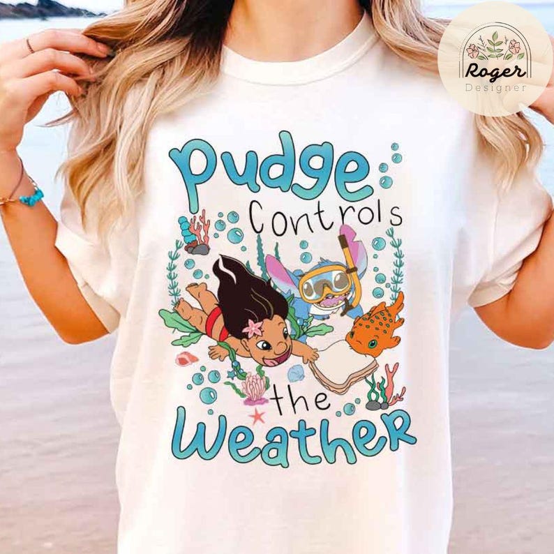 May include: White t-shirt featuring a colorful graphic with the text "Pudge Controls the Weather." The design includes cartoon characters, sea creatures, and bubbles in shades of blue, orange, and green. The shirt has a crew neck and short sleeves.