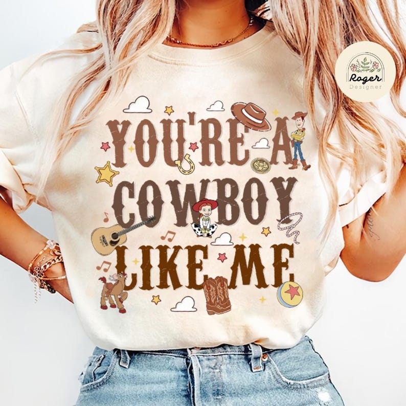 Może przedstawiać: Kremowy t-shirt z brązowym napisem "YOU'RE A COWBOY LIKE ME" otoczonym kolorowymi ilustracjami, takimi jak kowbojski kapelusz, gitara, lasso, koń, piłka, gwiazda oraz postacie z kresk&oacute;wek w graficznym projekcie o tematyce westernowej.