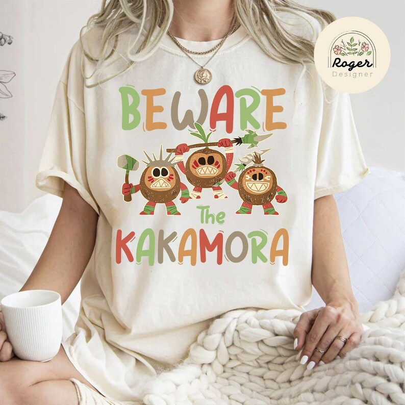 Puede incluir: Una camiseta blanca con el texto "BEWARE The KAKAMORA" en letras de colores. El texto est&aacute; rodeado por tres personajes de dibujos animados que parecen cocos con caras y extremidades.