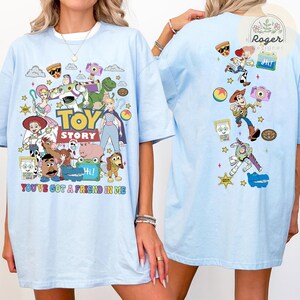 May include: A light blue t-shirt featuring a large Toy Story graphic on the front with characters like Woody, Buzz Lightyear, Jessie, and Rex, and the text You've Got A Friend In Me. The back displays a vertical arrangement of smaller character icons.
