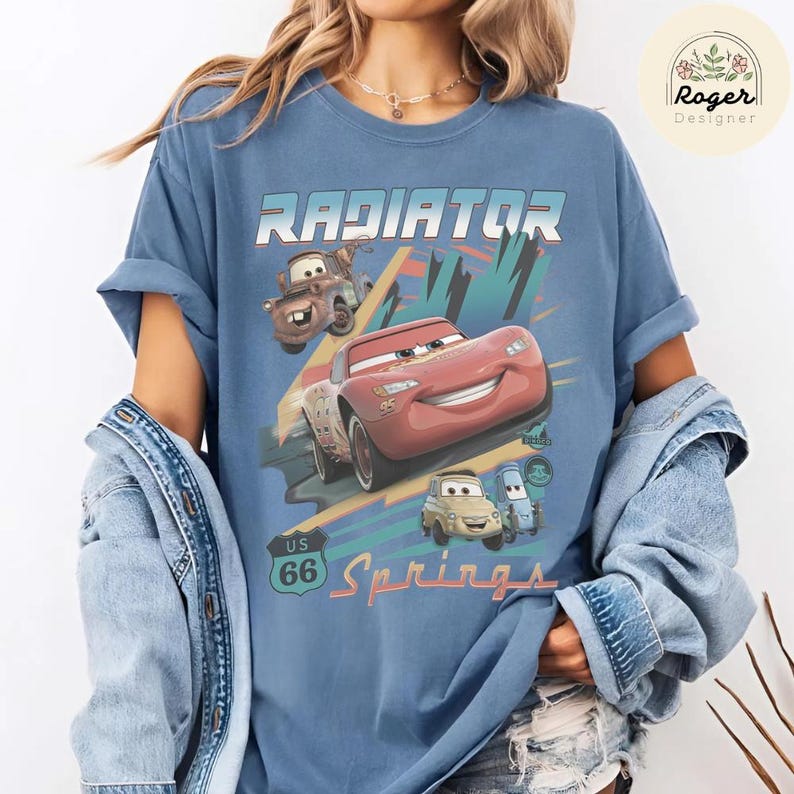 May include: A blue t-shirt featuring a graphic print of characters from the movie Cars, including Lightning McQueen, Mater, Luigi, and Guido. The design includes the text Radiator Springs and a US 66 road sign graphic in a vintage-inspired style.