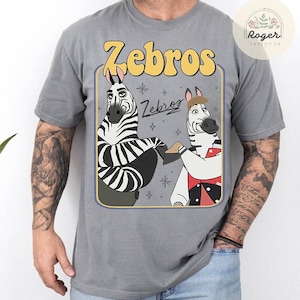 Pu&ograve; includere: T-shirt grigia con un design a cartoni animati di due zebre e la scritta "Zebros". Le zebre sono in una cornice rettangolare con un bordo giallo. La maglietta &egrave; realizzata in un materiale morbido e confortevole.