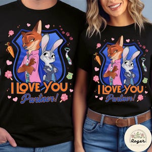 Pu&ograve; includere: T-shirt nere con un'illustrazione a cartoni animati di Nick Wilde e Judy Hopps di Zootropolis, con la scritta "I Love You, Partner!" in arancione e blu. Il design include carote, impronte di zampe e cuori.