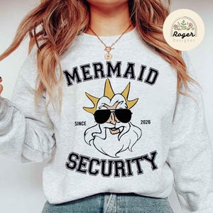 May include: A pale grey sweatshirt with the words "MERMAID SECURITY" in black, bold lettering. It features a cartoon illustration of a bearded figure wearing sunglasses. The text "SINCE 2026" is also visible.
