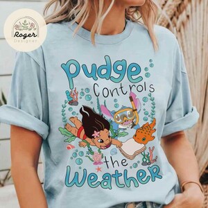 May include: Light blue t-shirt featuring a cartoon graphic with the text "Pudge Controls the Weather." The graphic includes characters and sea life. The shirt has rolled-up sleeves and a round neckline. The shirt has a logo in the upper left corner.
