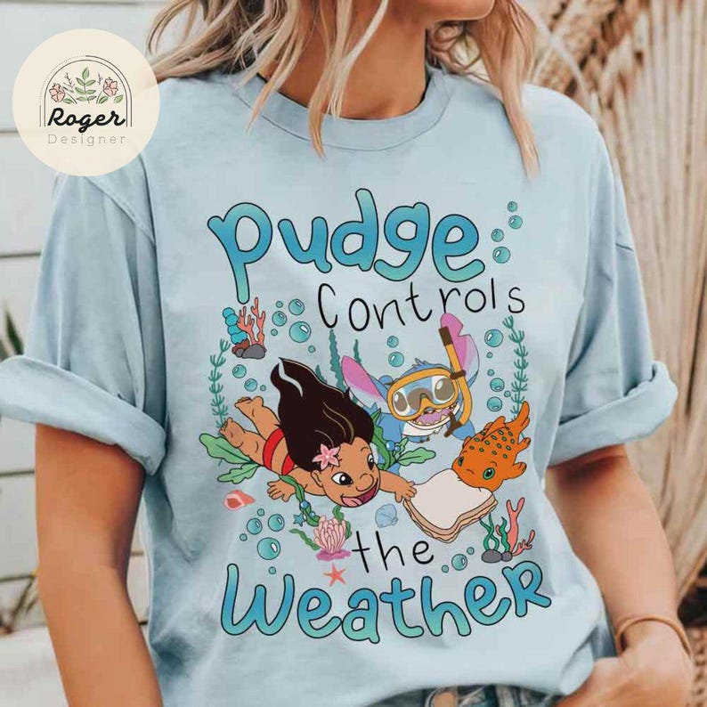 May include: Light blue t-shirt featuring a cartoon graphic with the text "Pudge Controls the Weather." The graphic includes characters and sea life. The shirt has rolled-up sleeves and a round neckline. The shirt has a logo in the upper left corner.
