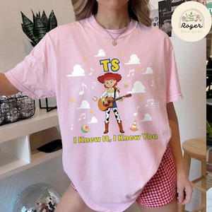 K&ouml;nnte beinhalten: Ein hellrosa T-Shirt mit einer Grafik einer Cartoon-Cowgirl, die eine akustische Gitarre spielt. Das Design enth&auml;lt den Text TS und I Knew It, I Knew You, umgeben von wei&szlig;en Wolken, Musiknoten, einem gelben Ball und einem orangefarbenen Pylon.