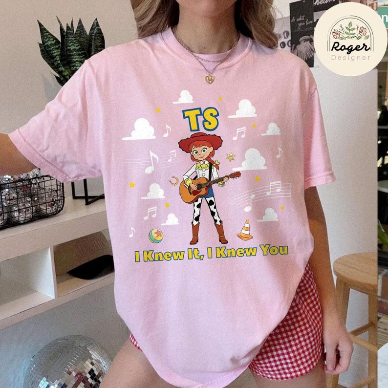 K&ouml;nnte beinhalten: Ein hellrosa T-Shirt mit einer Grafik einer Cartoon-Cowgirl, die eine akustische Gitarre spielt. Das Design enth&auml;lt den Text TS und I Knew It, I Knew You, umgeben von wei&szlig;en Wolken, Musiknoten, einem gelben Ball und einem orangefarbenen Pylon.
