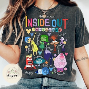 May include: A gray t-shirt with a colorful graphic of characters from the Disney Pixar movie Inside Out. The text "Inside Out" is in rainbow colors and the text "How are you feeling?" is in black. The characters are Joy, Sadness, Anger, Fear, Disgust, Bing Bong, and the other emotions.