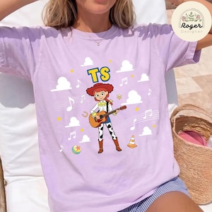 K&ouml;nnte beinhalten: Ein hellviolettes Kurzarm-T-Shirt mit einer Grafik einer Cartoon-Cowgirl, die eine akustische Gitarre spielt. Das Design enth&auml;lt den Text TS, wei&szlig;e Wolken, Musiknoten, einen gelben Stern, ein Hufeisen, einen bunten Ball und einen orangefarbenen Kegel.