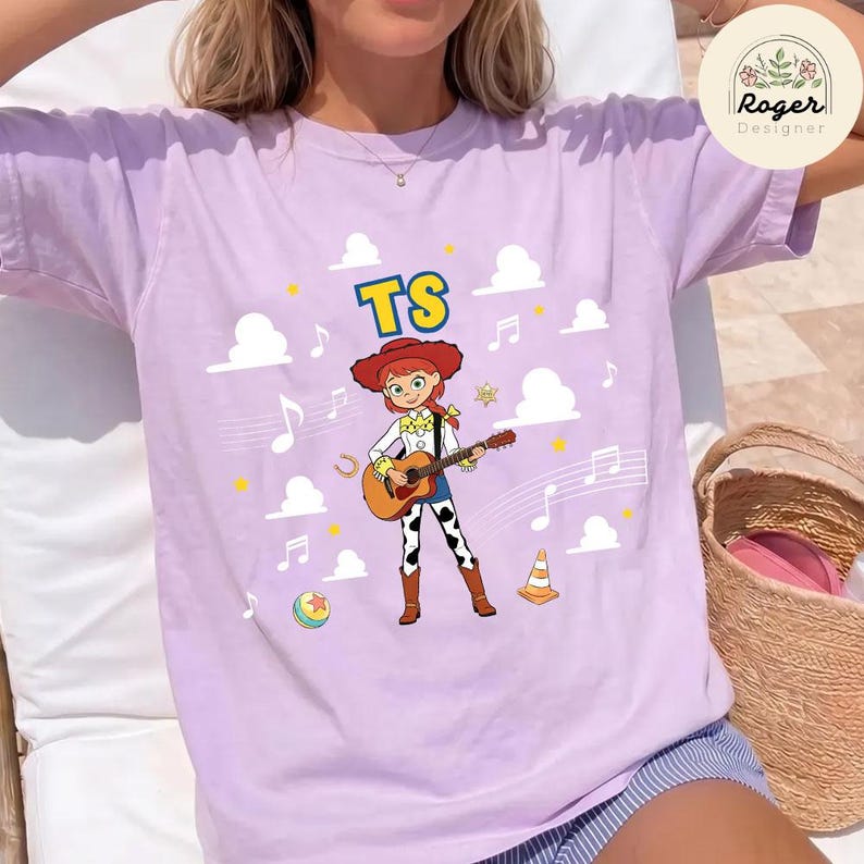 K&ouml;nnte beinhalten: Ein hellviolettes Kurzarm-T-Shirt mit einer Grafik einer Cartoon-Cowgirl, die eine akustische Gitarre spielt. Das Design enth&auml;lt den Text TS, wei&szlig;e Wolken, Musiknoten, einen gelben Stern, ein Hufeisen, einen bunten Ball und einen orangefarbenen Kegel.