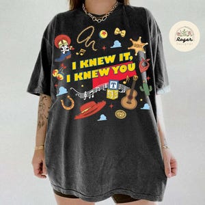 May include: A charcoal grey short-sleeve t-shirt featuring a graphic print with the text I KNEW IT, I KNEW YOU. The design includes illustrations of a cowboy boot, a lasso, a sheriff star, a red hat, a guitar, alphabet blocks, and musical notes.