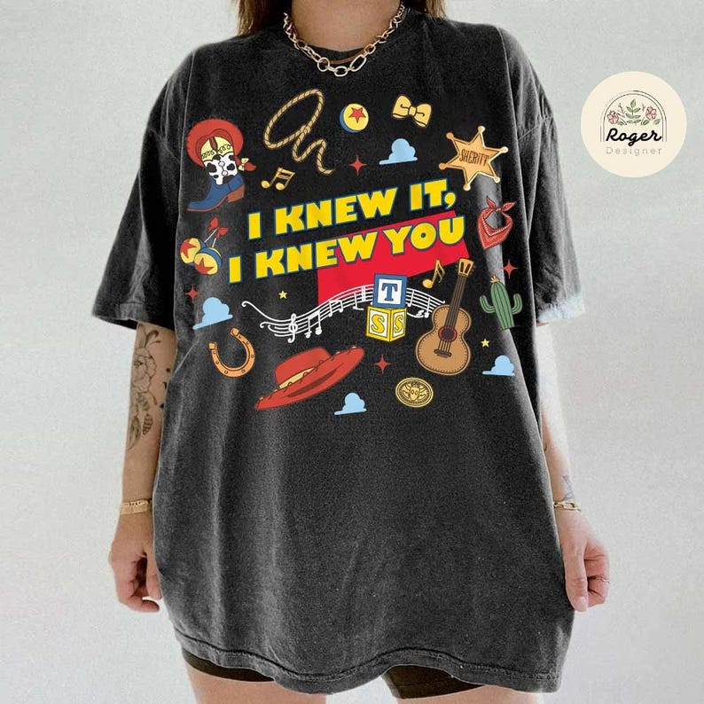 May include: A charcoal grey short-sleeve t-shirt featuring a graphic print with the text I KNEW IT, I KNEW YOU. The design includes illustrations of a cowboy boot, a lasso, a sheriff star, a red hat, a guitar, alphabet blocks, and musical notes.