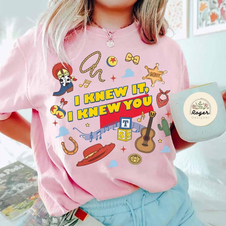 May include: A light pink t-shirt featuring the text I KNEW IT I KNEW YOU in yellow and white. The graphic includes a cowboy boot, lasso, sheriff badge, guitar, red hat, alphabet blocks, and musical notes. The shirt is paired with a light blue mug.