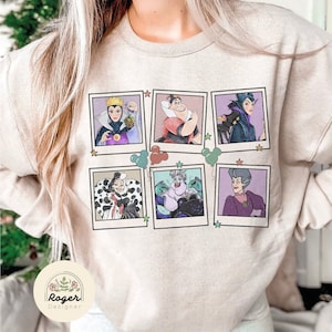 May include: A beige sweatshirt featuring a graphic print of six Disney villains in Polaroid frames. The villains are Maleficent, Cruella de Vil, Ursula, Evil Queen, Lady Tremaine, and Queen of Hearts. The print is surrounded by stars and balloons.