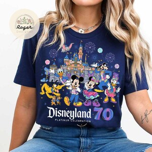K&ouml;nnte beinhalten: Ein marineblaues T-Shirt mit einem bunten Motiv von Mickey Mouse und seinen Freunden, die das 70-j&auml;hrige Jubil&auml;um von Disneyland feiern. Das Motiv zeigt Mickey Mouse, Minnie Mouse, Donald Duck, Goofy und Pluto. Der Text "Disneyland 70 Platinum Celebration" ist unter dem Motiv aufgedruckt.