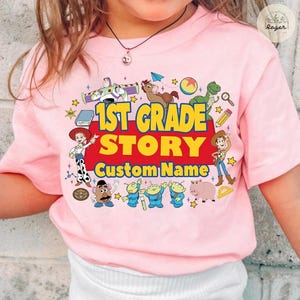 Pu&ograve; includere: T-shirt rosa chiaro con una grafica colorata. Il testo "1st Grade Story" e "Custom Name" &egrave; circondato da personaggi dei cartoni animati ed elementi scolastici. Un capo divertente e personalizzato.