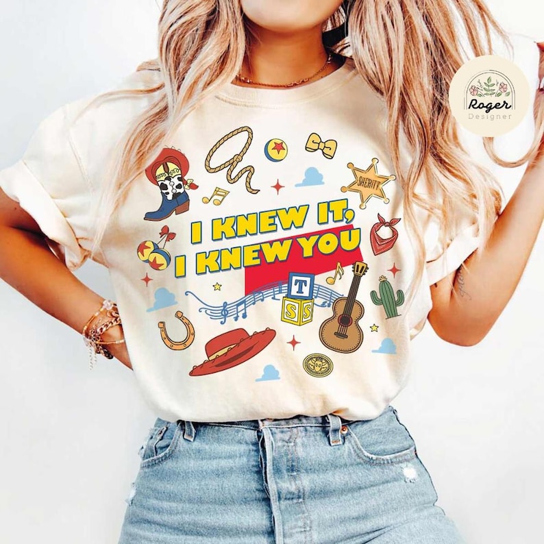 May include: A cream-coloured t-shirt featuring a colourful graphic print with the text I KNEW IT, I KNEW YOU. The design includes illustrations of a cowboy boot, a lasso, a sheriff star, a guitar, a cactus, a horseshoe, a hat, and alphabet blocks.