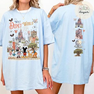May include: A light blue t-shirt featuring a large graphic on the front with the text Walt Disney World, depicting the castle, Epcot sphere, and various cartoon characters. The back of the shirt displays a vertical arrangement of park icons.