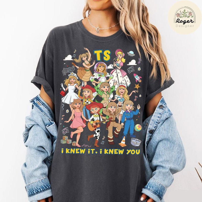 Op de afbeelding: Een antracietgrijs t-shirt met korte mouwen en een grafische print van meerdere stripfiguren in diverse outfits. De tekst "TS" staat bovenaan en de zin "I KNEW IT, I KNEW YOU" is in het geel onderaan het ontwerp gedrukt.